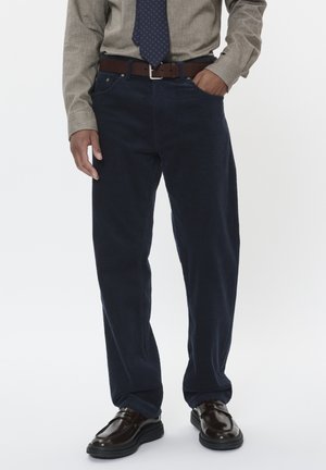 Marineblaue Corduroyhose mit geradem Schnitt, ausgestattet mit Seitentaschen und einem braunen Ledergürtel, kombiniert mit dunklen Slippern und einem hellen Hemd.