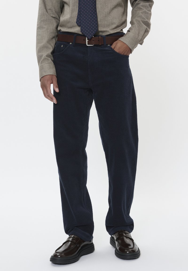 Pantaloni in corda blu navy con vestibilità dritta, dotati di tasche laterali e una cintura in pelle marrone, abbinati a mocassini scuri e una camicia chiara.
