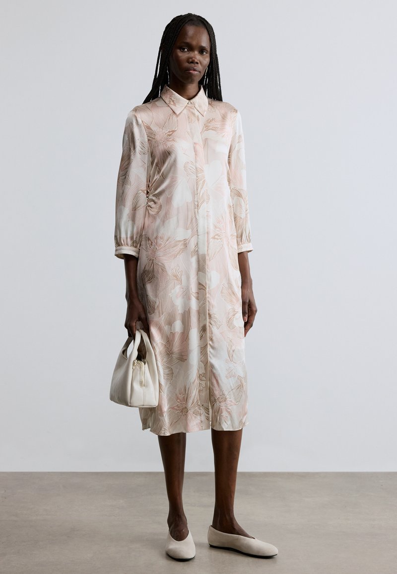 Φόρεμα ανοιχτού ροζ χρώματος με ανθισμένο μοτίβο, γιακά και μανίκια 3/4, κατασκευασμένο από μαλακό ύφασμα. Συνδυάζεται με λευκή τσάντα χειρός και μπεζ μπαλαρίνες.