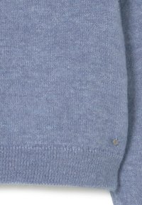 Tejido de suéter de punto azul suave con dobladillo acanalado y una pequeña etiqueta metálica redonda cerca del borde inferior.