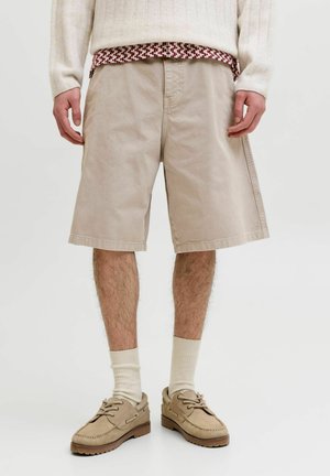Homme portant un short beige mi-genoux, un pull crème, des chaussettes crème et des chaussures décontractées beiges à lacets, debout sur un sol blanc.