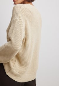 Femme portant un pull en grosse maille beige avec des manches longues et amples et un pantalon noir, sur un fond blanc uni.