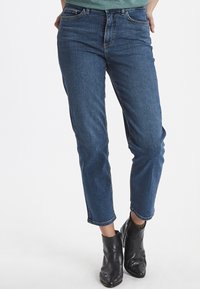 Jeans de mezclilla azul oscuro con corte recto, que cuentan con cinco bolsillos y costuras en contraste. Combinados con botas negras hasta el tobillo.