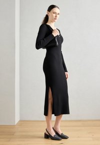 Robe noire à côtes avec manches longues, détails de boutons, fente latérale et design ajusté. Portée avec des chaussures à talons ouverts texturées noires.
