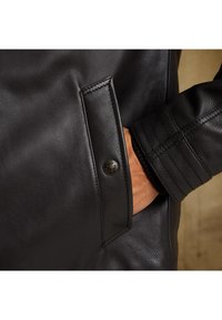 Čierna kožená bunda s textúrovaným povrchom, zipsovou kapsou a detailom na gombíku na manžete. Dizajn je priliehavý s rebrovanými akcentmi.