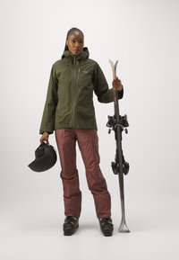Mørkegrønn vanntett jakke, burgunder skibukser, svart hjelm, holder ski. Teksturerte, funksjonelle materialer med hette og glidelås.