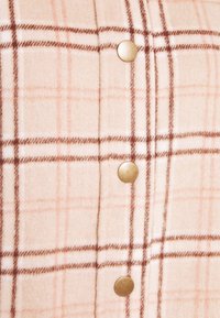 Tessuto a quadri in beige chiaro con linee pesca e marroni, caratterizzato da cinque bottoni tondi dorati lungo la chiusura. Materiale morbido e texturizzato.