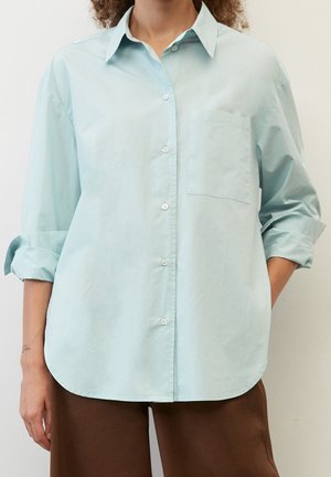 Persoon draagt een lichtblauw overhemd met opgerolde mouwen en een bruine broek, staand tegen een effen achtergrond.