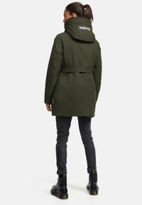 Veste imperméable vert olive avec un col haut, capuche réglable, et ceinture à la taille, associée à un pantalon slim gris foncé et des bottes noires.