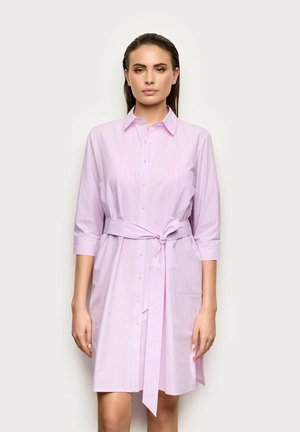 Femme portant une robe chemise rose clair boutonnée avec de fines rayures verticales et une ceinture nouée à la taille, debout devant un fond uni.