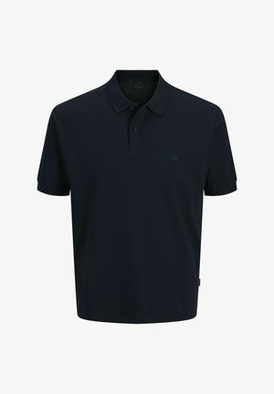 Jeune personne debout face à l'avant portant un polo noir, un short en jean retroussé, des chaussettes blanches et des baskets blanches sur un fond uni.