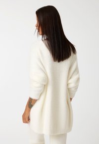 Maglione bianco morbido con maniche a tre quarti, caratterizzato da una vestibilità morbida e un'orlo posteriore più lungo. Texture liscia e dettagli di design minimal.