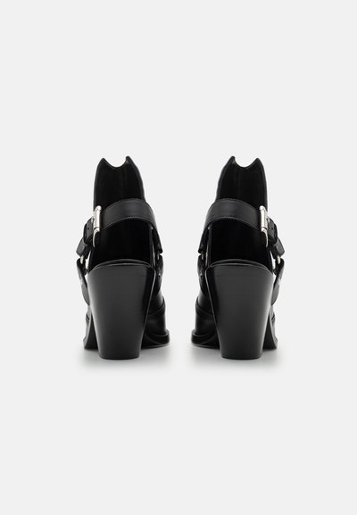 Black heeled shoes with a glossy finish, featuring a strap and buckle detail. Distinctive high back and thick, flared heel.