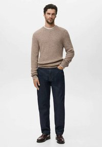 Man in een beige gebreide trui, donkere spijkerbroek en donkerbruine leren schoenen, staand met één hand in zijn zak tegen een effen achtergrond.
