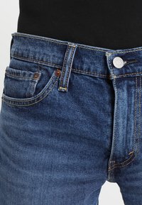 Mörkblå denimjeans med en klassisk femfickdesign, prydda med bronsnitar, gul sömnad och en metallknapplåsning.