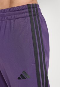 Lilla treningsbukser med svarte sidestriper, elastisk midje, teksturert stoff og en liten svart logo på underbenet.