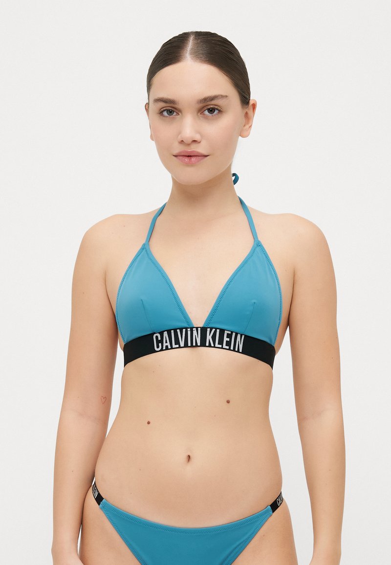 Teal triangeltopp med svart midjeband som har "CALVIN KLEIN"-logotyp. Materialet är glansigt med en åtsittande design och minimal hårdvara.