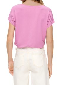 Top rosa a maniche corte con scollatura rotonda, realizzato in tessuto morbido. Il modello lo indossa con pantaloni bianchi a vita alta, che mostrano una vestibilità rilassata.