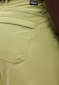 Artimos nuotraukos šviesiai alyvuogių žalios spalvos Patagonia kelnės, matomas užpakalinis kišenė ir virš jo esanti prekės ženklų etiketė.