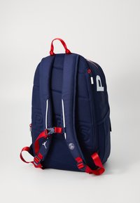 Navy blue backpack with padded straps, red accents, and logo details. Features two side zip pockets and a structured rectangular shape.