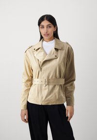 Giacca in satin beige con vita cincherata, zip frontale e ampi revers. Presenta accessori in argento e una vestibilità rilassata, indossata sopra una maglietta bianca.