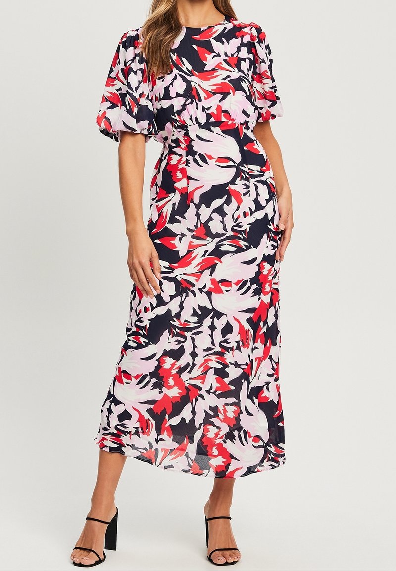Femme portant une robe midi fleurie avec manches bouffantes en rouge, noir et rose pâle, associée à des sandales noires à talons blocs et brides.