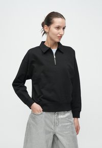 Μαύρο φούτερ με φερμουάρ και γιακά, με διακριτικό λογότυπο στο στήθος και άνετη εφαρμογή. Φορεμένο με μεταλλικό γκρι παντελόνι.