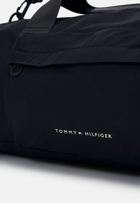 Černá taška přes rameno Tommy Hilfiger s přední kapsou na zip a horním držadlem, s logem značky v bílých, červených a modrých barvách.
