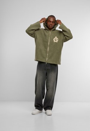 Olivgrüne Zip-Hoodie mit strukturiertem Finish, großer Kapuze und gesticktem Logo. Kombiniert mit lockeren grauen Jeans und weißen Sneakers.