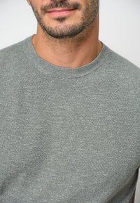 Grå stickad tröja med rund halsringning, med ett strukturerat mönster och ett mjukt, lätt tyg. Nära vy av halsringningsområdet.