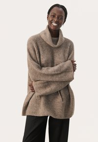 Hellbrauner Strickpullover, Rollkragen, lockere Passform, gerippte Textur, weite Ärmel, kombiniert mit dunklen Hosen, vor einem neutralen Hintergrund.