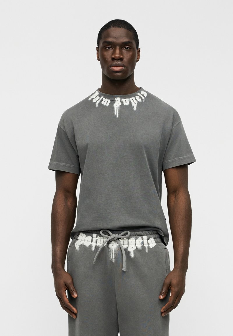Γκρι βαμβακερό μπλουζάκι με κοντά  μανίκια, με το λευκό γραφικό "Palm Angels" στο γιακά. Συνδυάζεται με αντίστοιχο παντελόνι φόρμας.