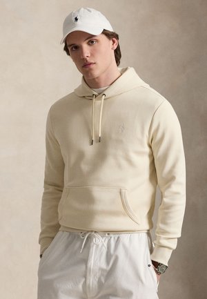Ung mand iført en cremefarvet hættetrøje, hvide bukser med snøre, hvid kasket med logo og et ur på venstre håndled, står foran en neutral baggrund.