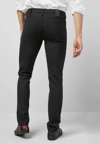 Zwarte slim-fit jeans met achterzakken, een gladde textuur en een donker merklabel op de taille. Gecombineerd met zwarte schoenen met rode en paarse accenten.