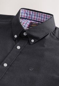 Chemise noire à boutons blancs, col intérieur à carreaux rouge, blanc et bleu, avec un petit logo couronne brodé sur la poitrine.