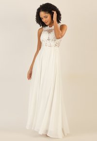 Weißes Kleid mit einem Spitzenoberteil, einem durchsichtigen Ausschnitt und einem fließenden Chiffonrock. Das Design umfasst kunstvolle Blumenmuster und eine sanfte Textur.