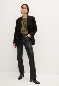 Blazer doppiopetto nero sopra una blusa a fantasia verde oliva, abbinato a pantaloni in pelle nera e stivali con tacco quadrato neri.