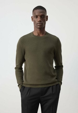 Man draagt een olijfgroene gestructureerde trui en zwarte broek, staat met handen in de zakken tegen een effen lichte achtergrond.