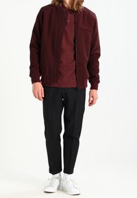 Man som bär en vinröd zip-up jacka och polo-tröja, svarta byxor och vita sneakers, stående mot en enkel vit bakgrund.