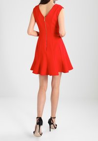 Czerwona sukienka bez rękawów z rozkloszowanym dołem, z dekoltem w literę V na plecach oraz złotym zamkiem z tyłu. Na nogach znajdują się czarne sandały na wysokim obcasie.