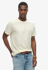 Cremegul t-shirt med korte ærmer lavet af bomuld, med rund hals og et lille logo patchet på venstre ærme. Båret med blå denim jeans.
