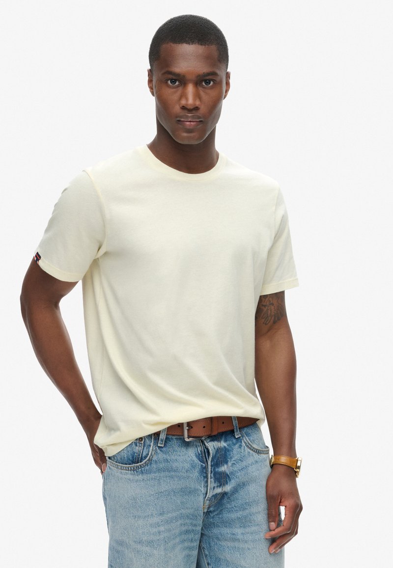 Cremegul t-shirt med korte ærmer lavet af bomuld, med rund hals og et lille logo patchet på venstre ærme. Båret med blå denim jeans.
