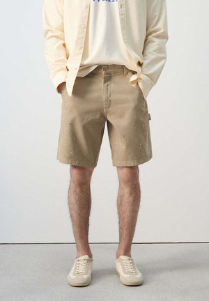 Mężczyzna ubrany w beżowe szorty poplamione farbą, kremowe sneakersy, białą koszulkę i jasnobeżową koszulę na guziki stoi z rękami w kieszeniach.