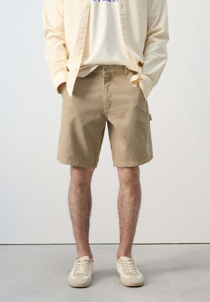 Hombre con pantalones cortos beige salpicados de pintura, zapatillas de color crema, camiseta blanca y camisa beige claro abotonada, de pie con las manos en los bolsillos.
