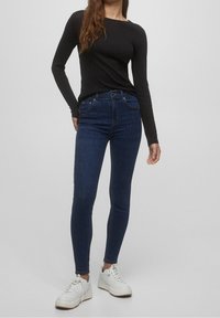 Jeans skinny azul oscuro con cintura alta y bolsillos frontales, combinados con una camiseta de manga larga negra y zapatillas blancas. Textura suave.