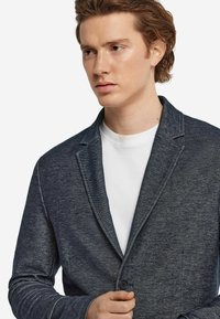 Marinblå cardigan i en texturerad tyg, med sjalkrage, enkel knäppning, öppen fram och sidofickor.