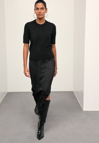 Top nero a maniche corte in maglia abbinato a una gonna nera in raso elegante con rifiniture in pizzo. Stivali neri in pelle al ginocchio completano il look.