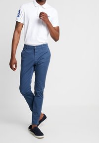 Vit piké med blå logotyp och detaljer, tillsammans med blå slim-fit byxor. Modellen bär marinblå slip-on skor. Snyggt tyg och skräddarsydd passform.