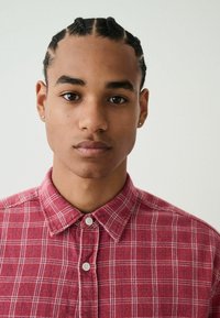 Rotes kariertes Hemd mit Knöpfen aus strukturiertem Stoff, mit Kragen und weißen Knöpfen. Das Modell trägt Zöpfe und hat einen neutralen Gesichtsausdruck.