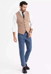Beige Weste mit Reverskragen, fünf Knöpfen und vorderen Taschen, kombiniert mit einem weißen Hemd und blauen Jeans. Schwarze Schuhe runden den Look ab.